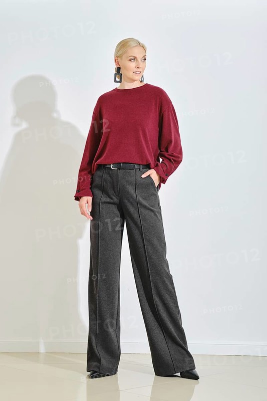 A fashion model stands confidently in a minimalist studio, wearing a maroon sweater and high-waisted black trousers. Bright daylight illuminates the space, enhancing her stylish look