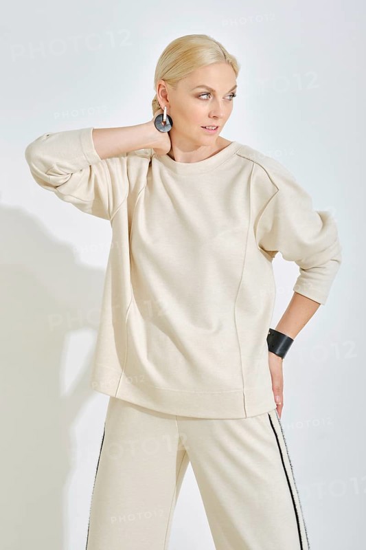 A woman showcases a light-toned casual outfit featuring a relaxed sweatshirt and stylish pants. She strikes a confident pose, highlighting her accessories against a clean white backdrop