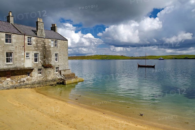 Old stone cottage on quiet loch shore with sandy beach and passing boat under cloudy sky, Murder in Shetland crime series, Viking ship, Up Helly A, Lerwick, Shetland Islands, Scotland, United Kingdom