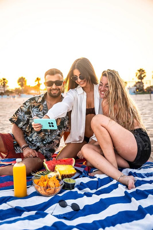 Three friends are taking a selfie with smartphone during a picnic at the beach, enjoying snacks and drinks