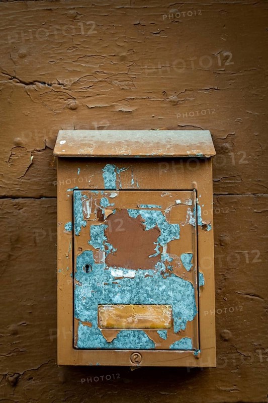 A rustic mailbox is mounted on a weathered brown wall, featuring peeling paint and faded colors. The blue paint reveals its age, while the wooden backdrop shows texture and wear. France
