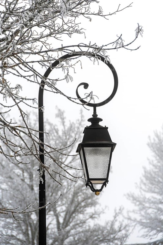 A decorative street lantern hangs from an ornate pole, surrounded by trees covered in frost. Aubrac, France