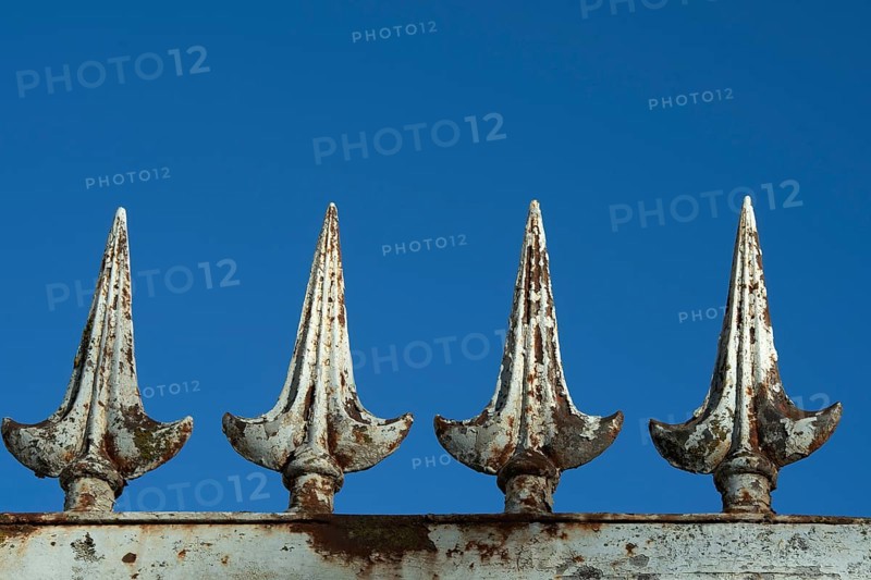 Four rusted metal spikes rise prominently against a vivid blue sky, showcasing weathered details that highlight their ornamental design and aged texture. France