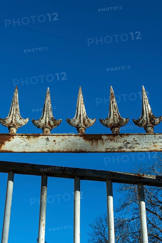 A weathered wrought iron fence stands tall under a bright blue sky. The fence has pointed spikes, showcasing signs of rust and age, adding character to the setting. France