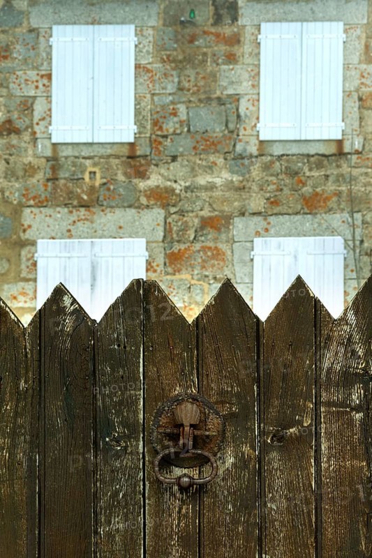 A weathered wooden fence stands in the foreground, with a charming stone house featuring white shutters in the background. Occitanie. France