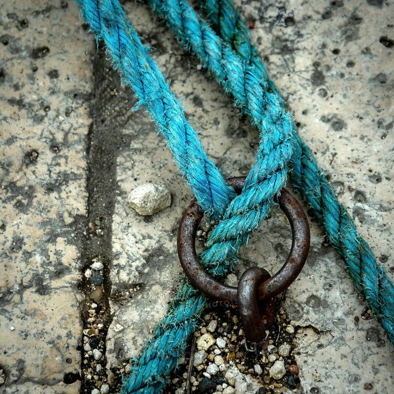 A close-up view reveals a vibrant blue rope intricately intertwined with a metal ring, resting on a textured and worn surface scattered with debris, highlighting the contrast in materials
