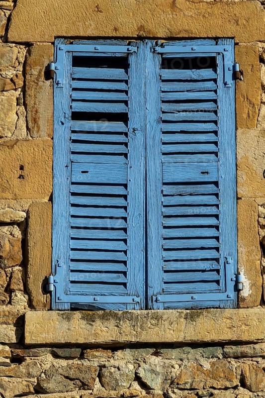 Bright blue shutters adorn the weathered stone wall of a traditional building in a picturesque village. Sunlight reflects off the vibrant color, creating a cheerful atmosphere. France