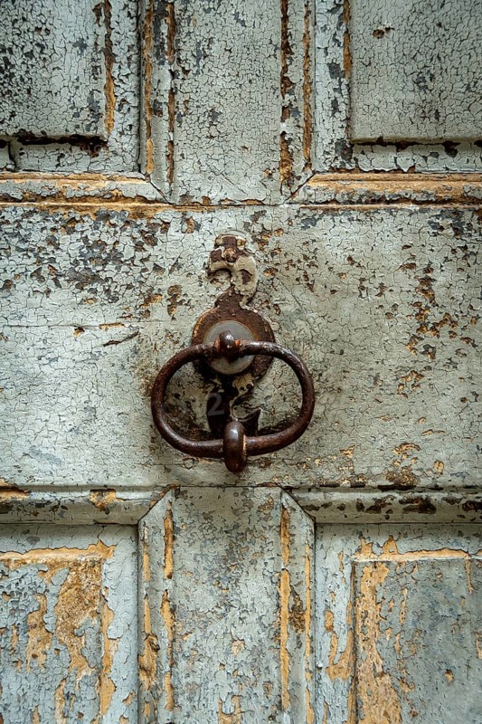 A close-up of a weathered wooden door surface reveals peeling paint and a vintage metal knocker. The texture tells a story of age and traditional craftsmanship