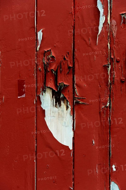 A close-up view captures the peeling red paint on a wooden surface, showcasing intriguing textures and patterns formed over time. The wear reflects exposure to the elements