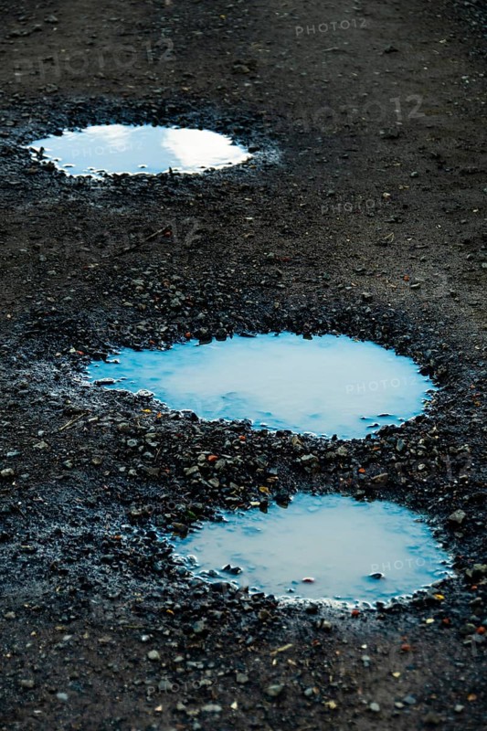 Three puddles on a gravel path mirror the overcast sky, highlighting the damp texture of the ground and creating a peaceful outdoor atmosphere after rainfall