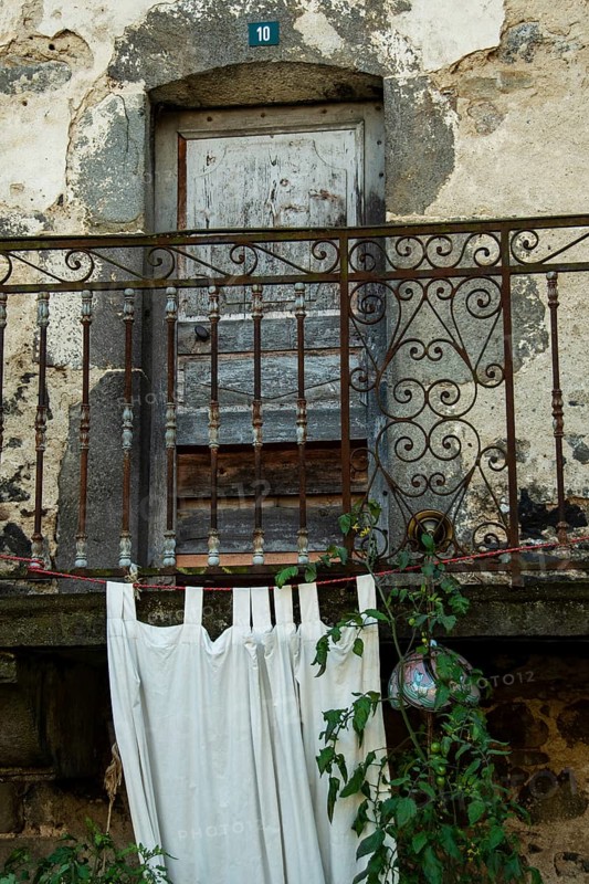 Located in an antique stone structure, this rustic balcony features an ornate railing, flowing white fabric, and climbing vines, evoking a sense of forgotten history and architectural beauty. France
