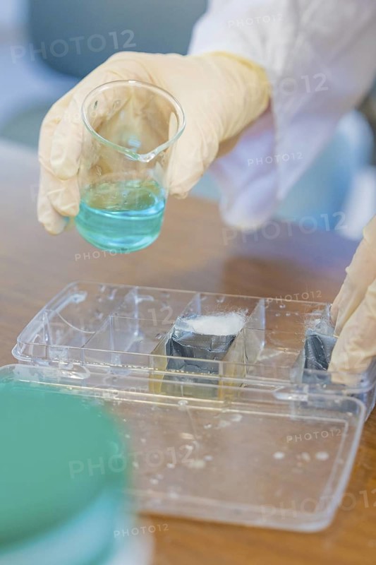 Person in gloves holding a beaker with blue liquid over a tub, Battery Research, Nagold, Germany