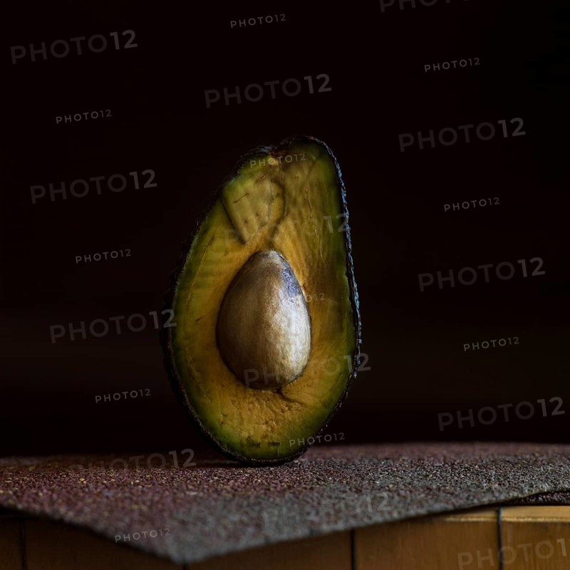 A half avocado is placed on a rough, textured surface, highlighting its smooth, creamy interior and prominent seed. The low lighting creates a rich, alluring atmosphere around the fruit