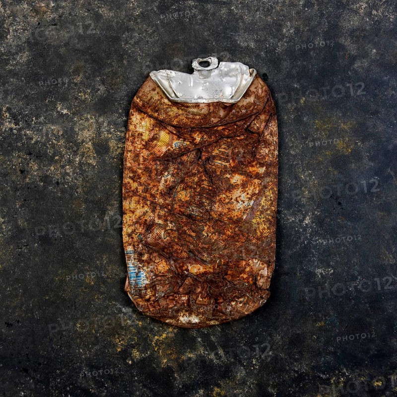 A rusty beverage can rests on a dark, textured concrete surface, highlighting urban litter and its impact on the environment over time. The can shows significant signs of corrosion and deterioration