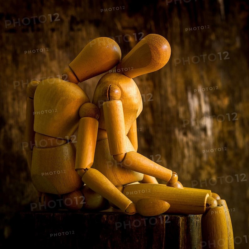 A wooden mannequin is positioned on a rough wooden surface, looking contemplative against a textured, dimly lit background. The warm tones enhance the somber mood of the scene