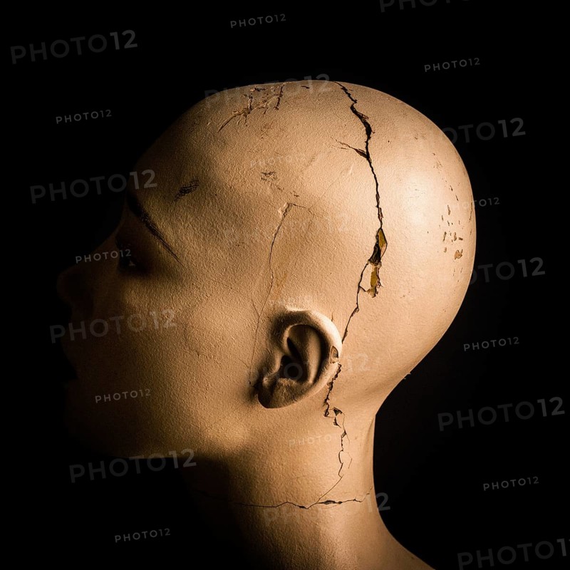 A head of a broken woman dummy is positioned in profile. The damaged surface reveals cracks and marks against a stark black background, emphasizing its worn features