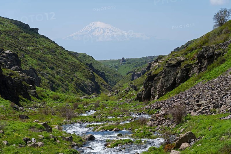 A river runs through a rocky landscape with a snow-covered mountain in the distance, Our Lady Church with Ararat in the background, Vahramashen Church, Vahramashen Surp Astvatsatsin, Amberd Gorge, Aragazotn Province, Armenia