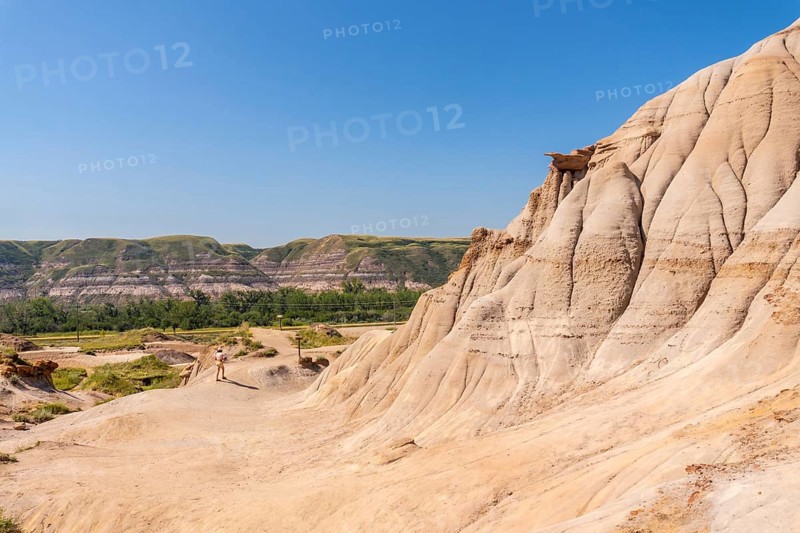 Tourist walking through the arid badlands of drumheller, alberta, surrounded by unique hoodoo formations under a clear blue sky, highlighting the region's natural beauty