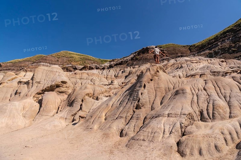 Tourist standing on a hoodoo rock formation, exploring the unique badlands landscape of drumheller, alberta, under a clear blue sky, enjoying the vastness and beauty of nature