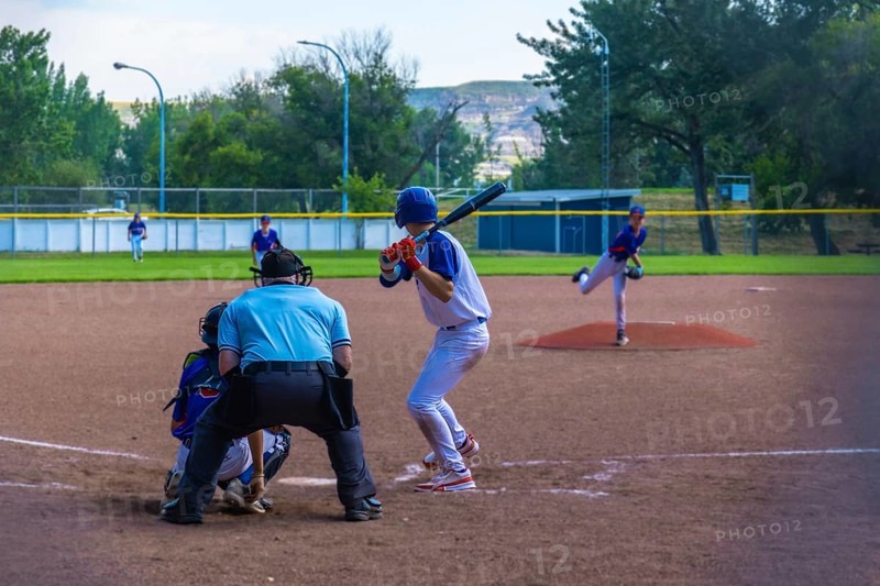 Teenage baseball batter waiting for the pitch while the pitcher is throwing the ball during a sunny summer game, with the umpire and the catcher behind him