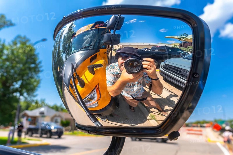 Photographer is taking a selfie reflected in the mirror of a truck, with a yellow school bus and parked cars in the background, in drumheller, alberta, on a sunny day