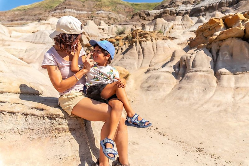 Mother holds her son on her lap, applying sunscreen to his nose during a sunny day exploring the unique hoodoo formations of drumheller, alberta