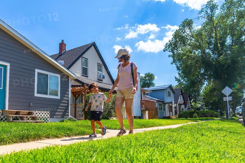 Mother and son walking hand in hand on a sunny day along a sidewalk in a peaceful drumheller neighborhood, surrounded by green lawns and charming homes