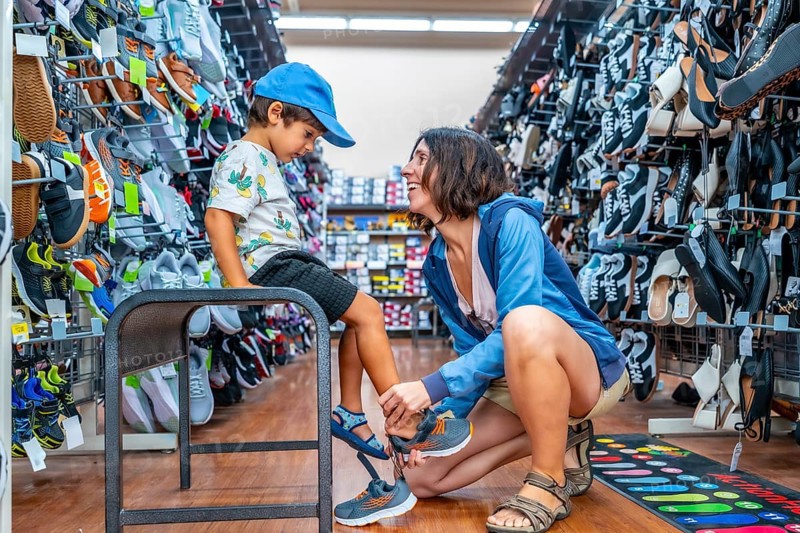 Mother smiling while assisting her son in trying on new shoes at a shoe store, surrounded by a wide selection of footwear, enjoying quality time together during their shopping experience