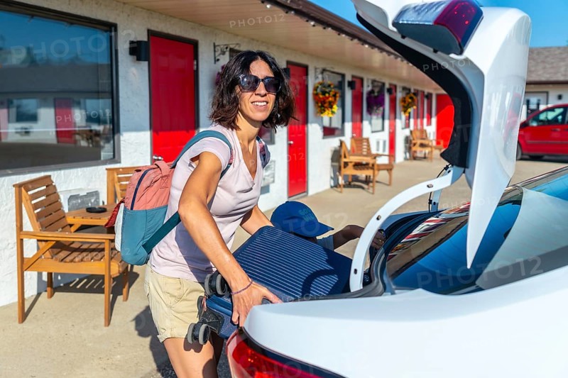 Female tourist wearing sunglasses and a backpack is loading her luggage into the trunk of her car outside a motel on a bright, sunny summer day, ready for an exciting adventure