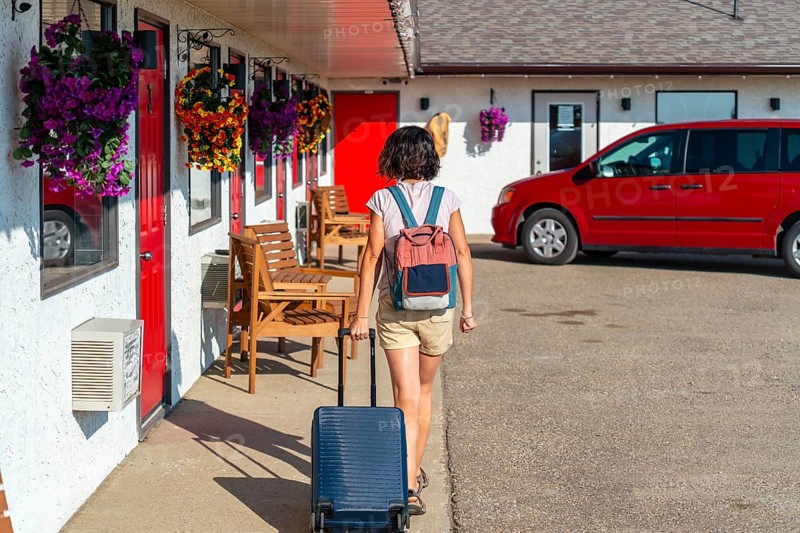 Female tourist walking towards her room at a roadside motel, pulling rolling luggage and wearing a backpack under the warm sun, enjoying the journey on a beautiful summer day