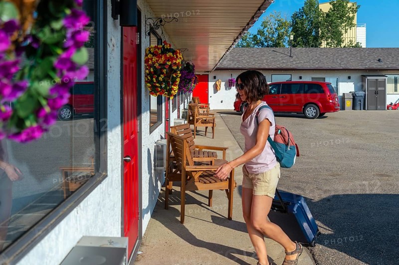 Female tourist is walking to her motel room pulling her wheeled suitcase, carrying a backpack and passing by some wooden chairs on a sunny summer day