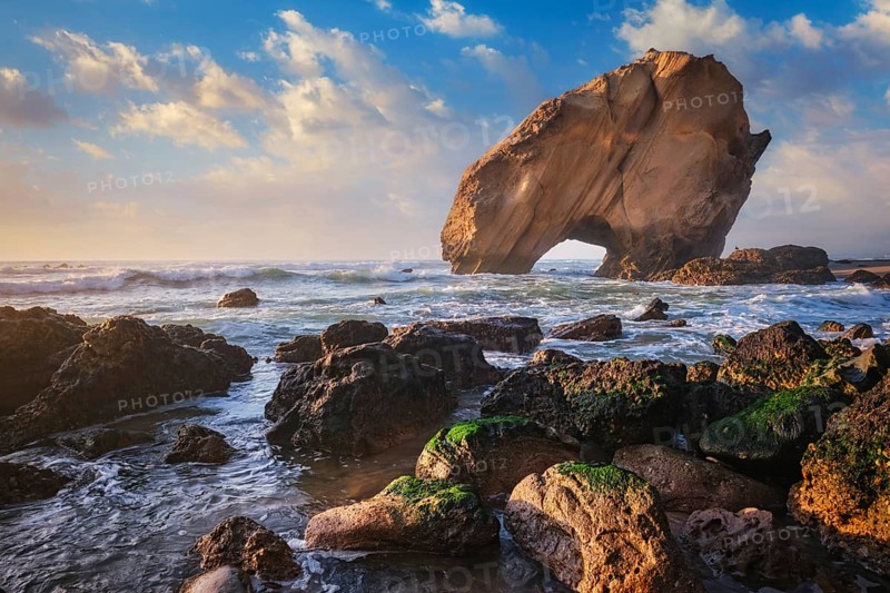 Penedo do Guincho, a large boulder rock arch at Praia da Santa Cruz, Portugal, with ocean waves and sandy beach on sunset