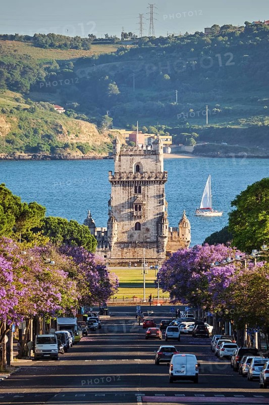 Scenic view of Belem Tower in Lisbon, Portugal, seen over a street with blooming purple jacaranda flower trees street with tourist sailboat on the Tagus River. Portugal