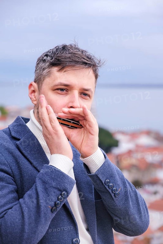Man musician in a blue blazer and white sweater playing blues on a harmonica with eyes closed, standing outdoors in city street