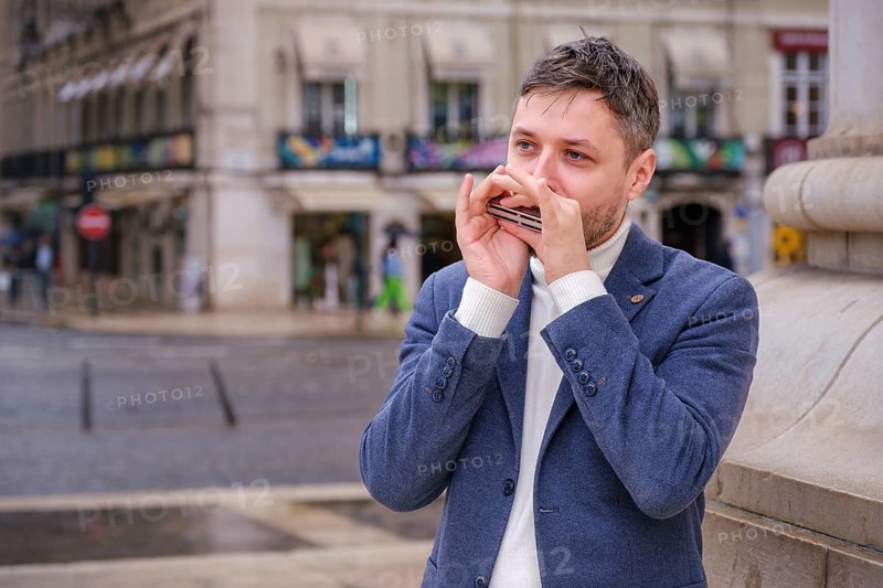 Man musician in a blue blazer and white sweater playing blues on a harmonica with eyes closed, standing outdoors in Lisbon city street, Portugal