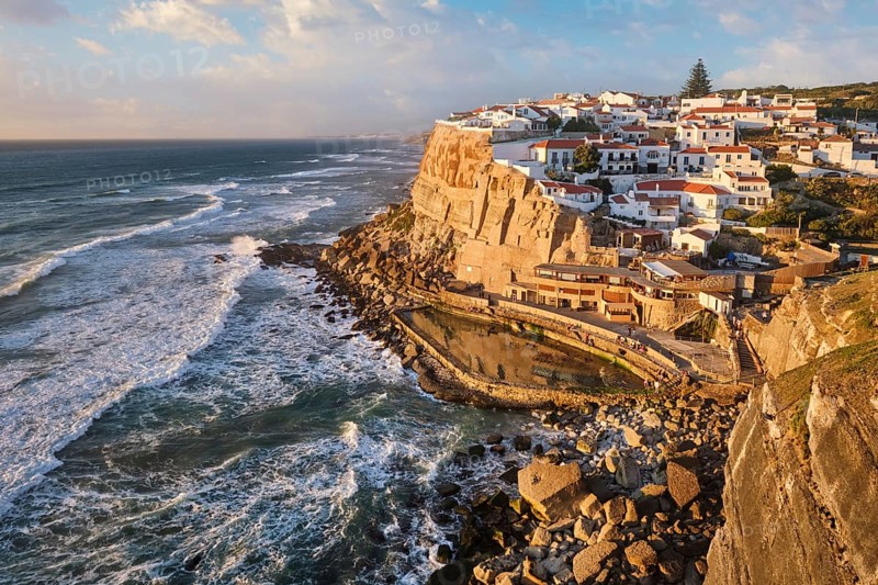 Charming Azenhas do Mar, a picturesque fishing village perched on cliffs. The sun sets over the Atlantic, casting warm hues on the whitewashed buildings and rocky coastline. Portugal