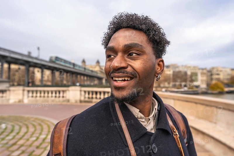 Happy black man smiling and looking away while strolling across paris bir hakeim bridge with a metro train passing, enjoying an urban citybreak by the seine