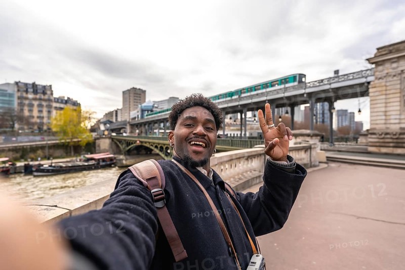 Happy african american man tourist taking a selfie portrait on a bridge over the seine river in paris, france, with the elevated metro passing by in the background