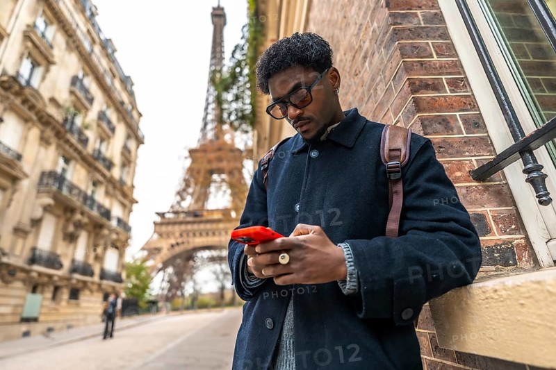 Young black man in paris leaning against a building, focusing on his smartphone, representing travel, technology, and urban modern connectivity in a famous european city