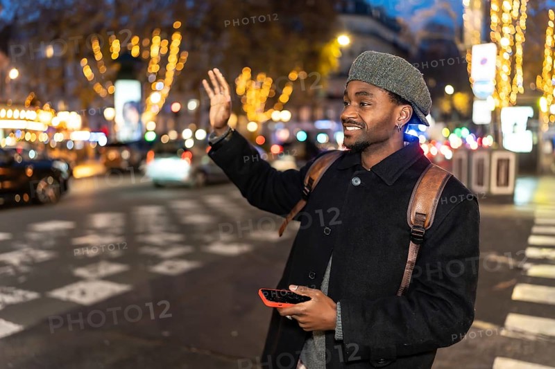 Young man smiling while hailing a taxi on a vibrant city street at night, holding a smartphone and wearing a backpack and hat, surrounded by blurred evening lights