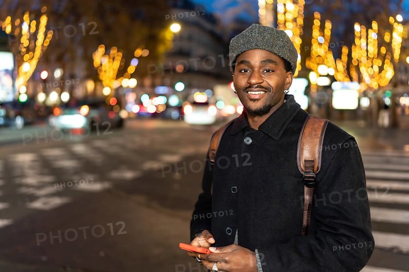 Young black man smiling in an urban setting at night, holding a mobile phone and standing on a city street with blurred car lights and decorated trees in the background