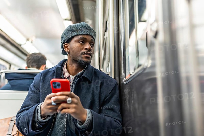 Young black man seated on subway, dressed for winter, holding smartphone and gazing out the window while commuting through the city, thoughtful and connected