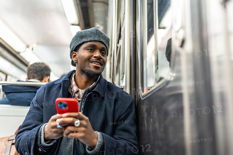 Young black man in beret and jacket rides the subway holding a red smartphone, smiling and looking out the window while commuting through the city, browsing and enjoying the ride