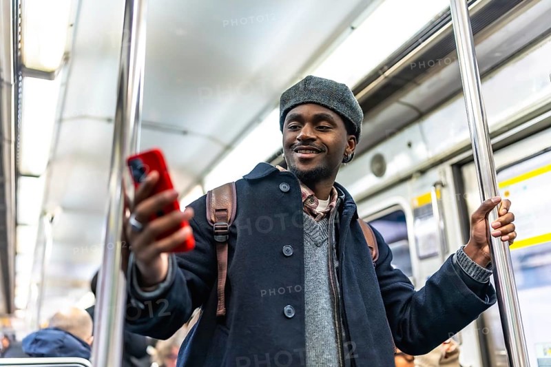 Young black man with a backpack holding onto a pole, smiling while enjoying social media on his red smartphone inside a moving public transport metro train