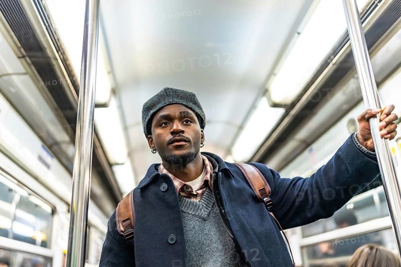 Young black man in winter coat and hat stands holding a subway pole on a crowded metro, looking away pensively during a daily commute, urban travel and routine