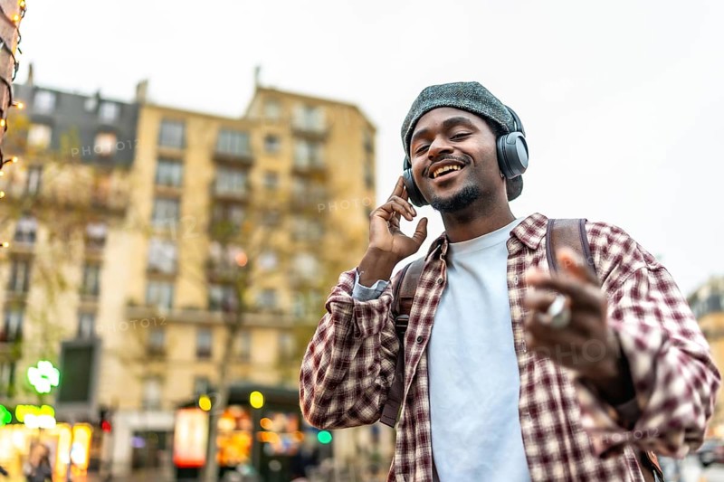 Young man in a beret and headphones smiling and dancing while walking through a vibrant urban street, enjoying music and carefree city life with confident, joyful energy