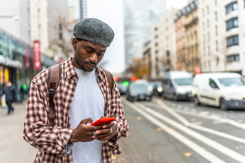 Young black man standing on an urban street, wearing a plaid shirt and a cap, engrossed in using his mobile phone, communicating or navigating in a busy city environment