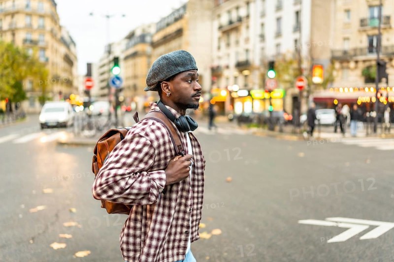 Young black man standing on a city street at a crosswalk, wearing a hat and backpack with headphones around his neck, looking right while waiting to cross