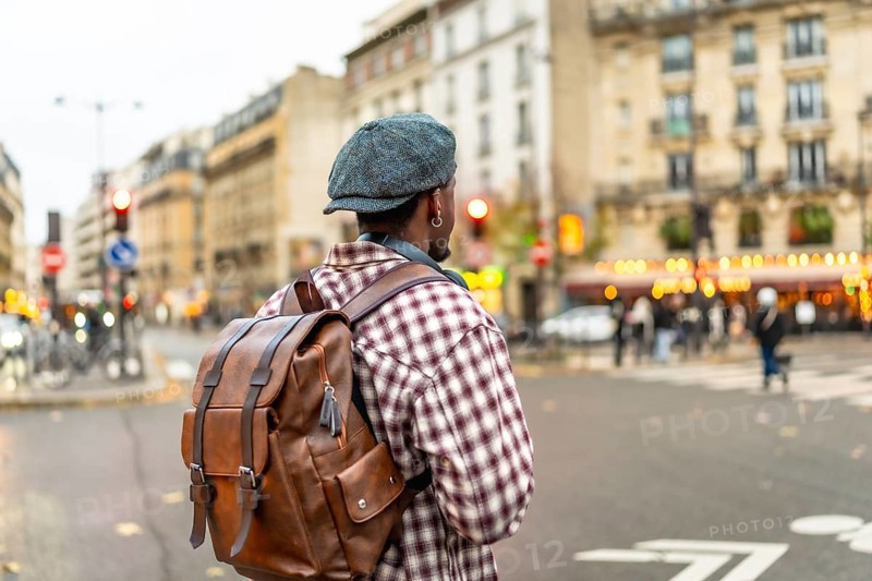 Young man wearing a cap and brown backpack stands on the curb of an urban street, looking across the busy intersection, ready to continue his journey through the city
