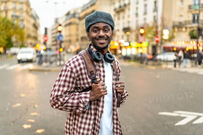 Young black man with headphones wearing a plaid shirt and backpack, standing confidently in a bustling european city street, enjoying urban exploration and a relaxed travel lifestyle